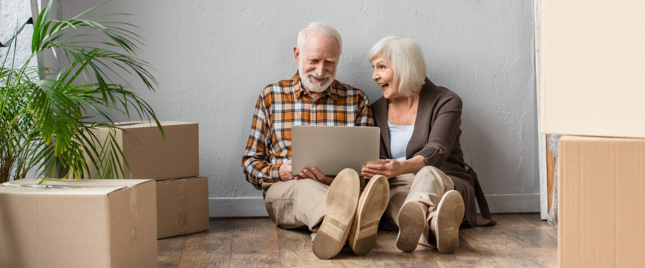 A senior man and woman sit on a hardwood floor against the wall, laughing and using a computer, with packed cardboard boxes surrounding them.