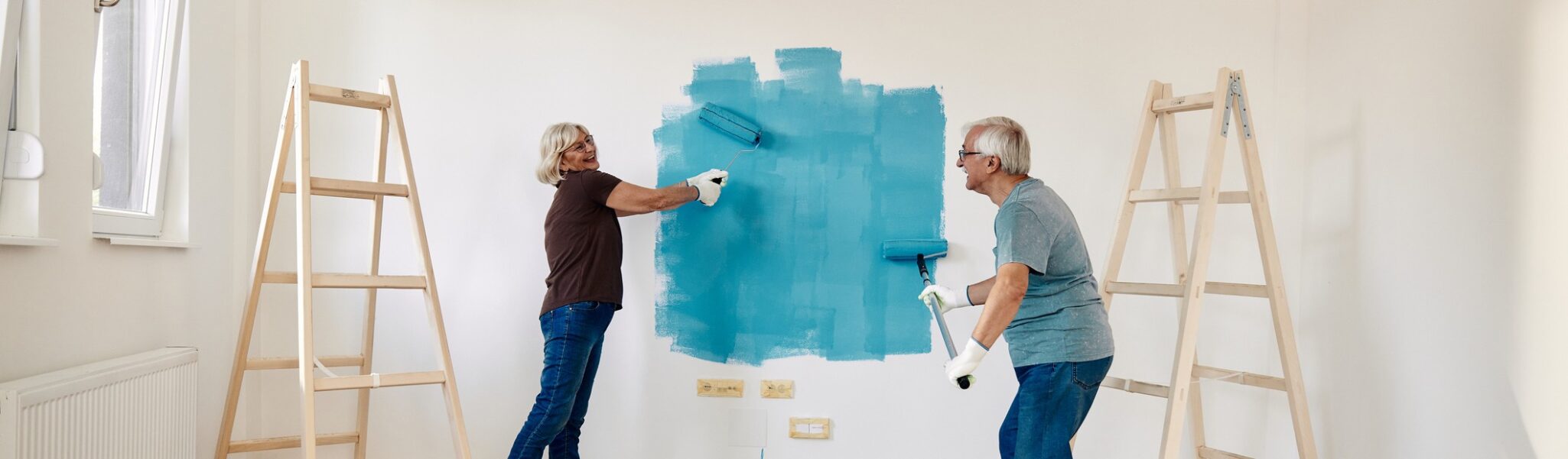 A happy senior couple paints a wall blue with rollers.