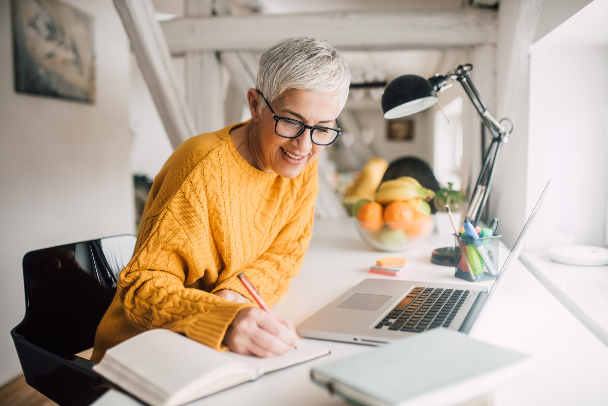 A senior woman wearing a yellow sweater, writing in a notebook while at an at home office desk